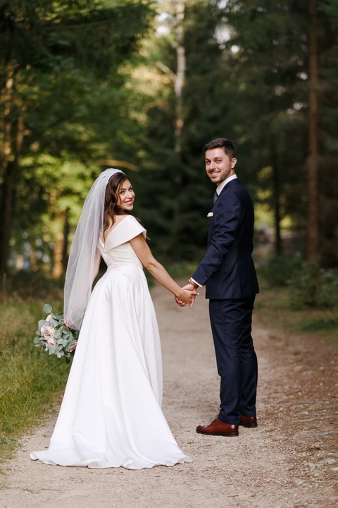 Novomanželé se drží za ruce a přes rameno koukají na fotografa, Harrachov