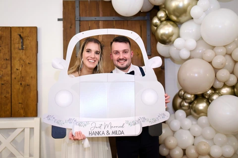 Novomanželé drží papírové autíčko a smějí se na fotografa