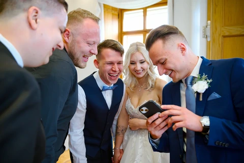 Novomanželé se svatebčany se smějí a koukají do mobilu, Štábní domek