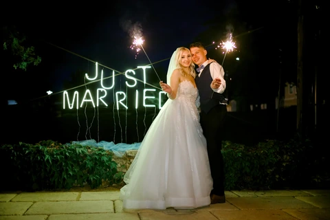 Novomanželé s prskavkami stojí u nápisu Just Married.