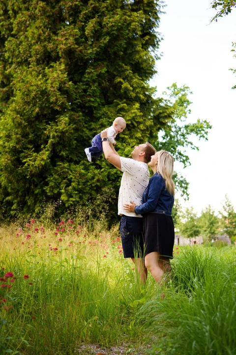 Tatínek zvedá syna do vzduchu a maminka přihlíží, rodinné focení v parku.